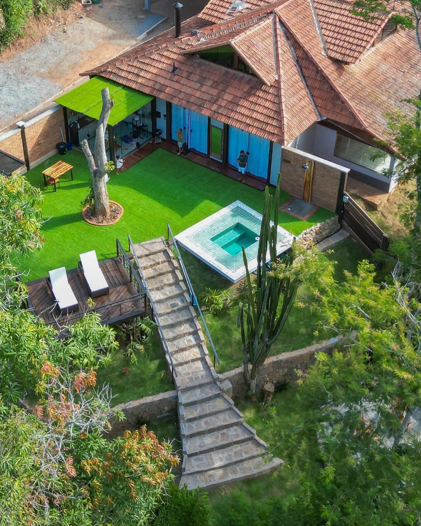 Vista de cima do bangalô: telhados de telha vermelha, gramado com tronco escultórico, piscina de pastilha verde, deck com espreguiçadeiras e a escada de pedra descendo pelo terreno