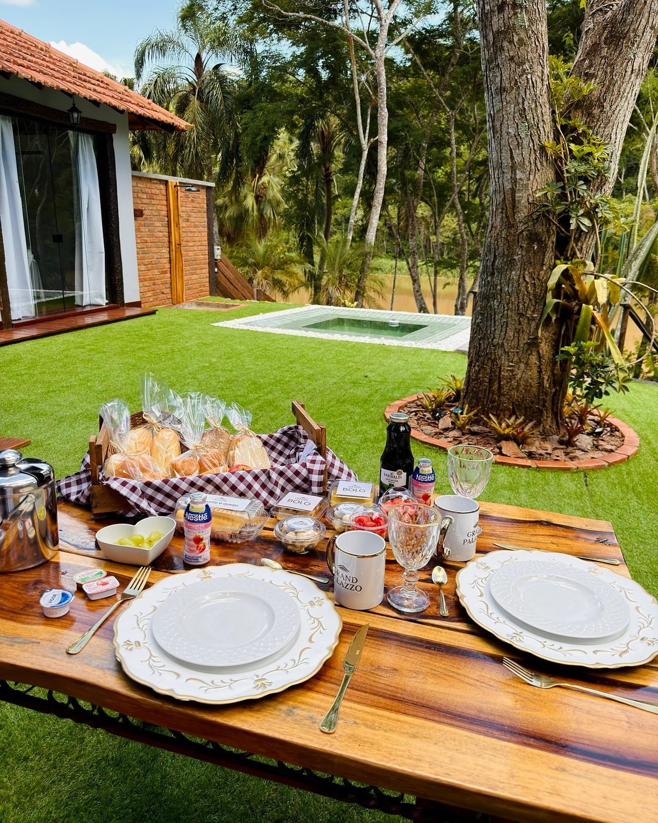Mesa de café da manhã posta no gramado, com pães e frutas, tendo a piscina e o bangalô ao fundo