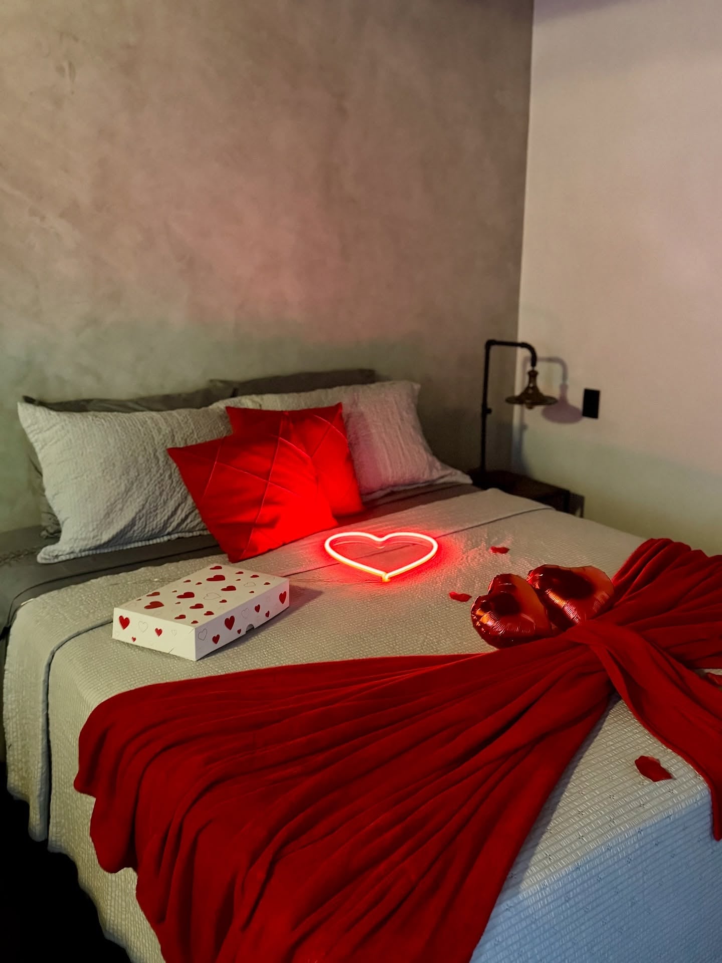 Cama de casal com colcha branca, almofadas vermelhas e decoração romântica com coração iluminado