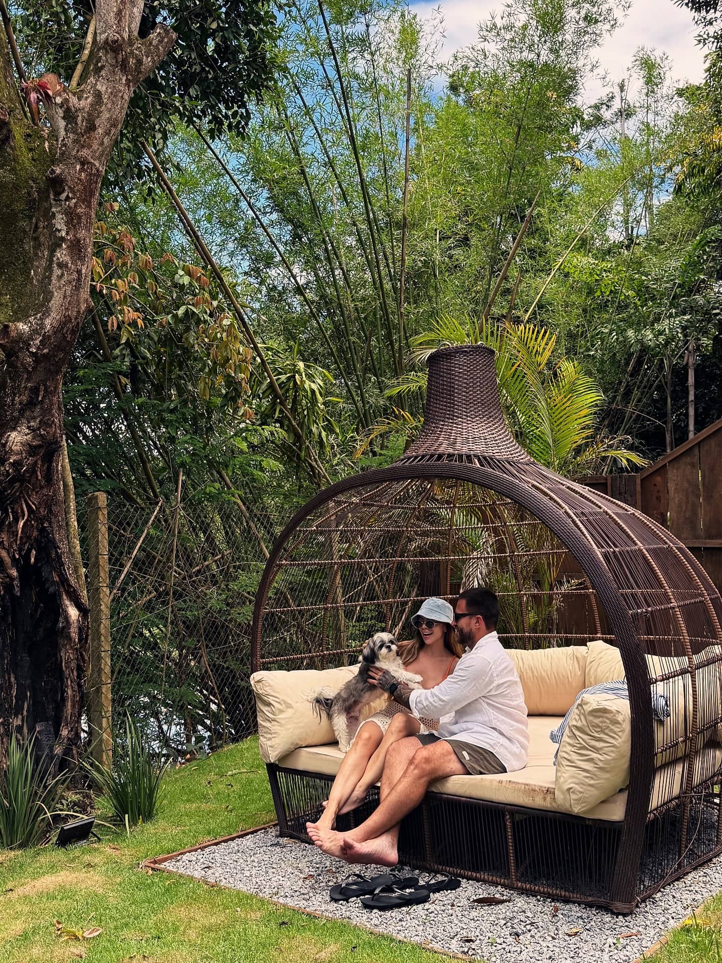 Daybed redondo de fibra com almofadas claras sob a copa das árvores, no jardim do bangalô
