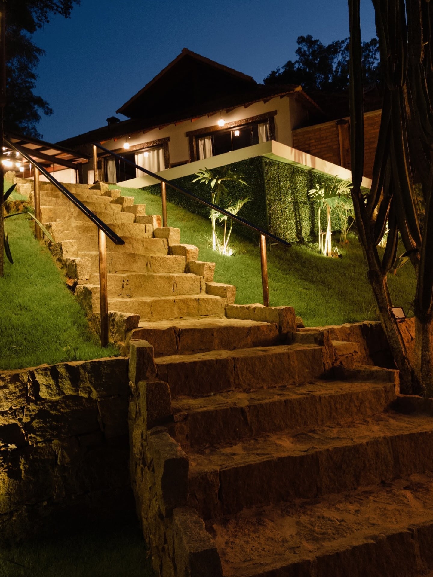 Escada de pedra iluminada subindo até o bangalô à noite, com as luzes quentes do beiral acesas