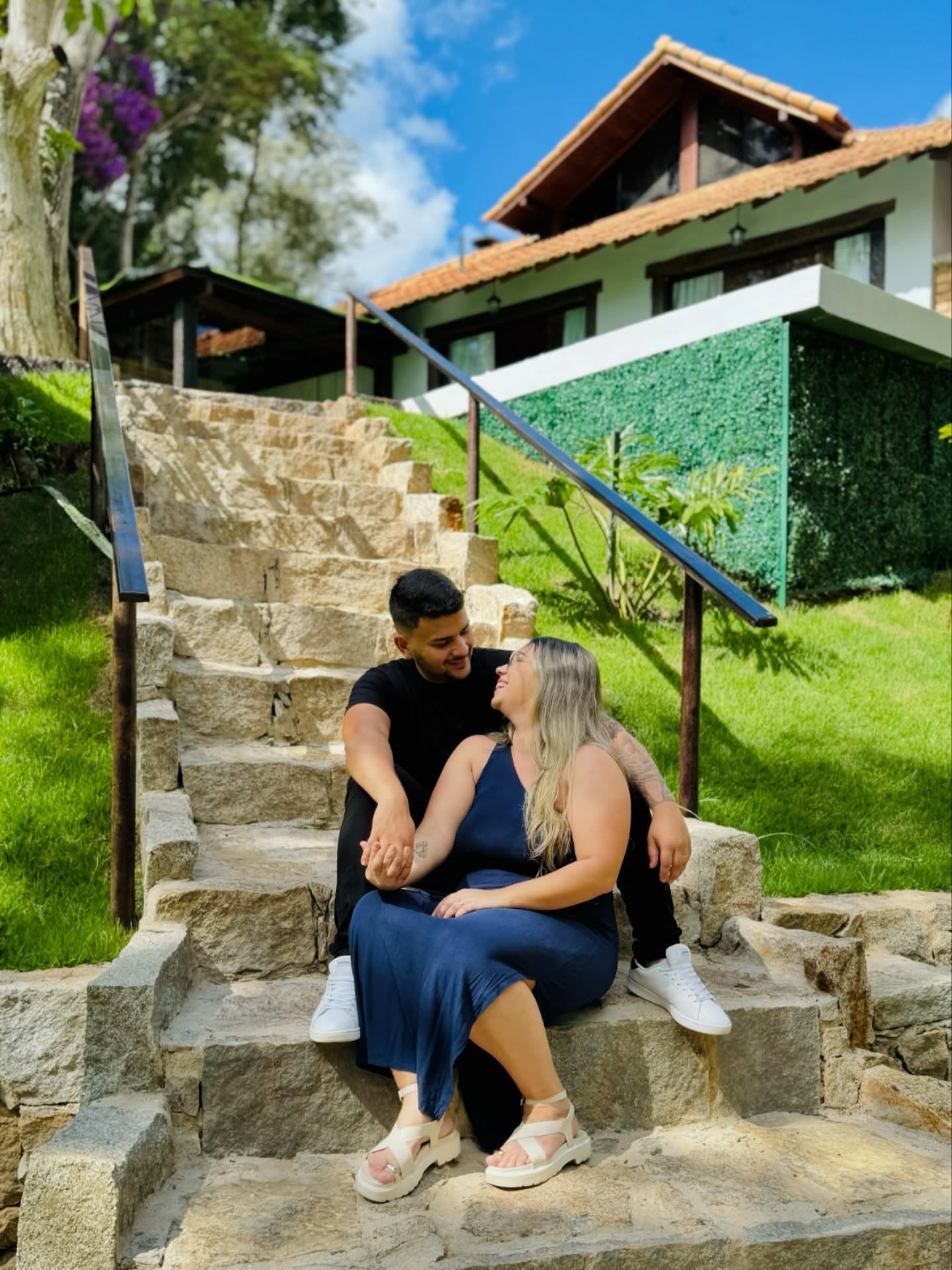 Escada de pedra com corrimão subindo pelo jardim até o bangalô, cercada de grama e muro verde
