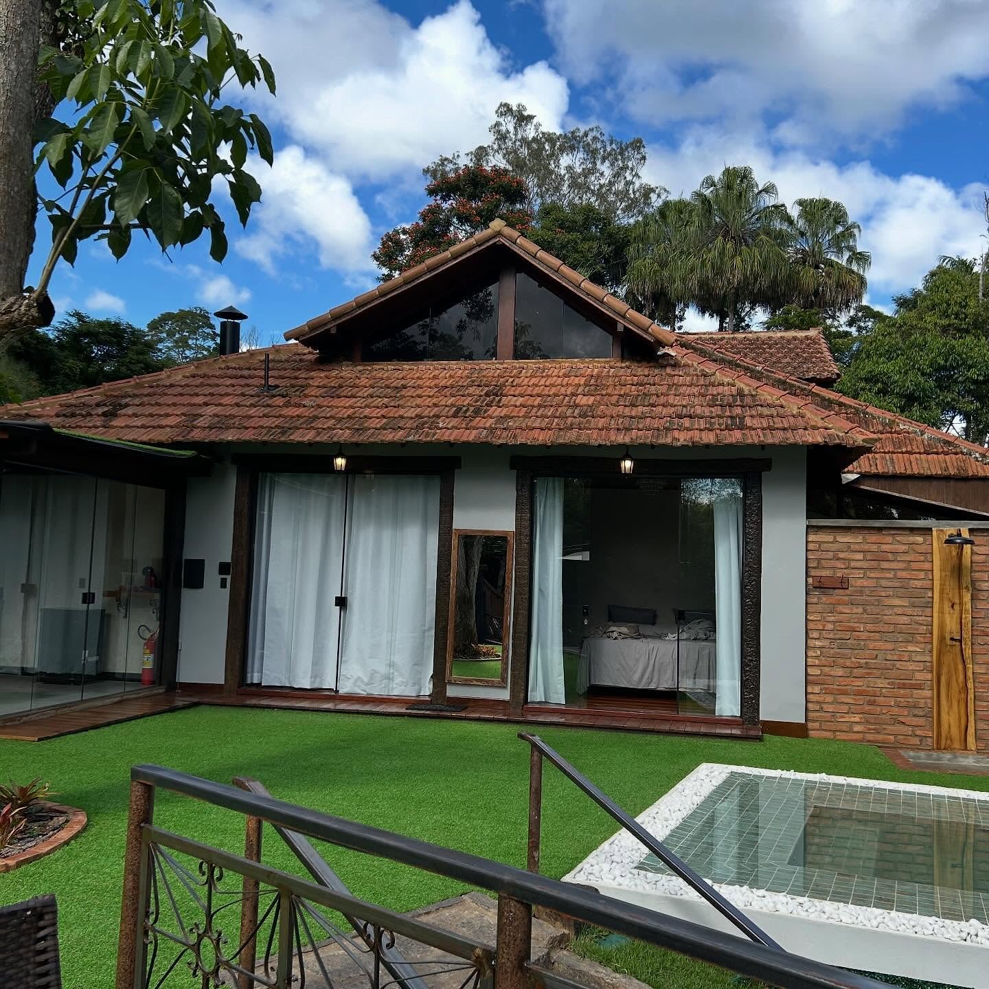 Fachada do bangalô em dia de sol, com portas de vidro e cortinas claras abrindo para o gramado e a borda da piscina