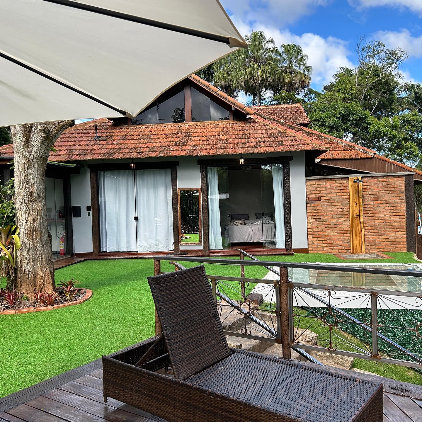 Fachada do bangalô de telha e madeira com espreguiçadeira e ombrelone sobre o gramado, entre árvores altas