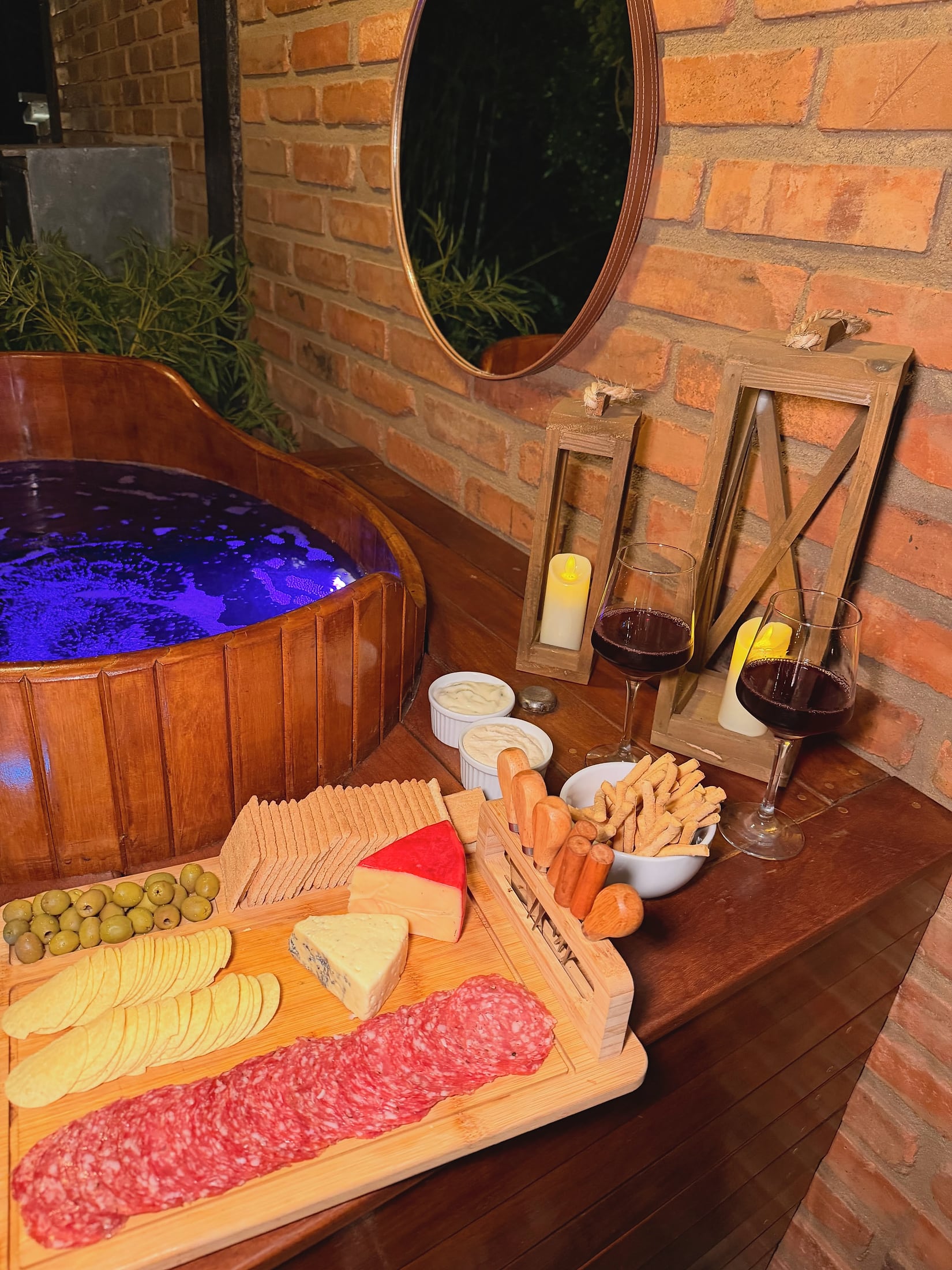 Hidromassagem redonda de madeira com água iluminada em azul, ao lado de tábua de frios e vinho sobre a bancada