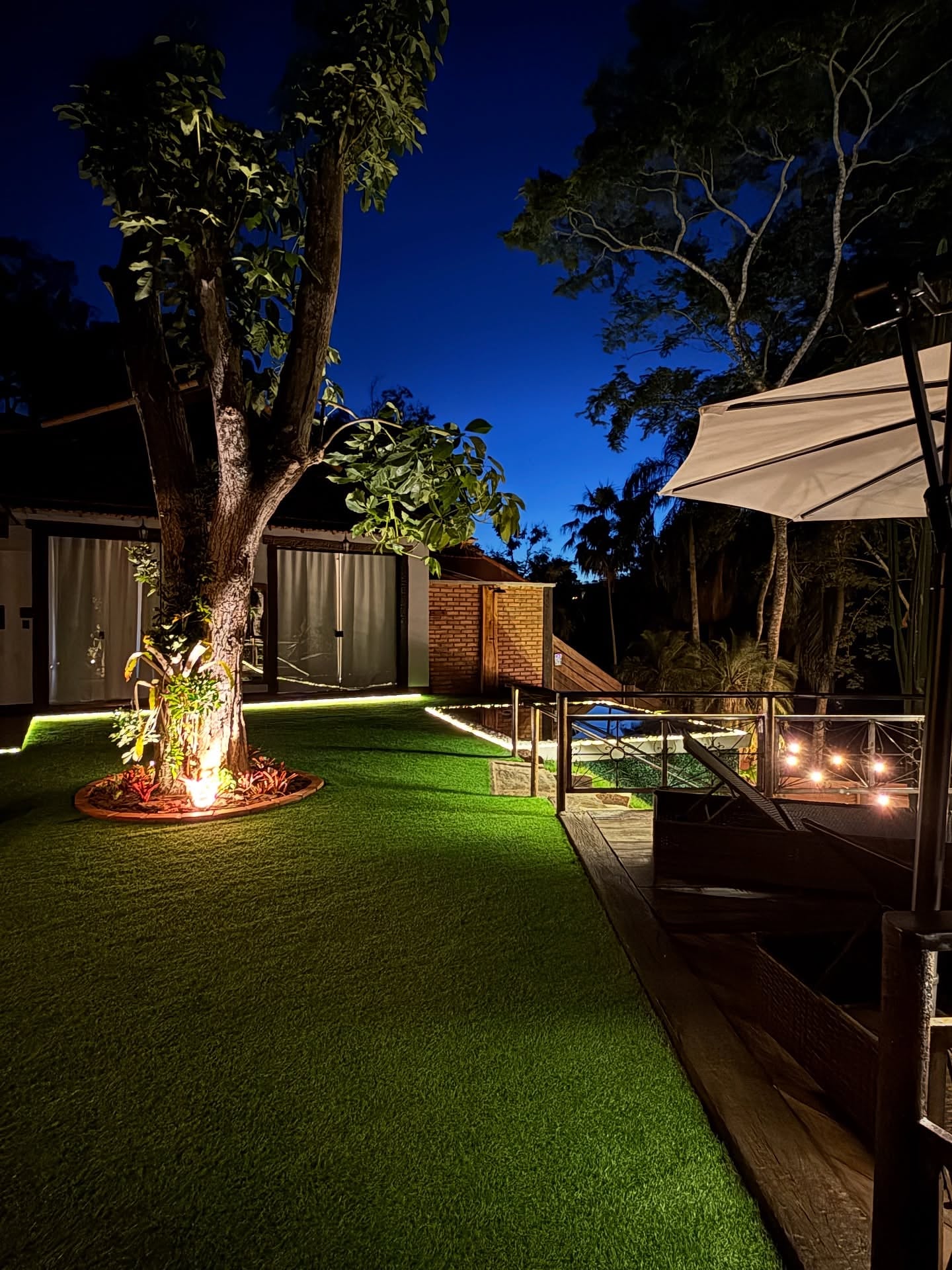 Jardim à noite com a árvore iluminada, o deck de madeira aceso por velas e o ombrelone aberto ao lado da piscina