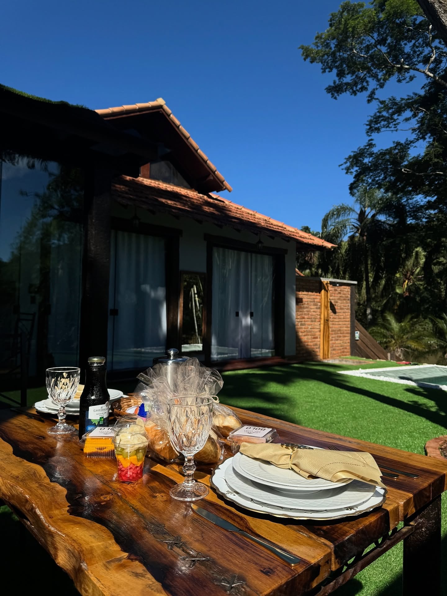 Mesa de madeira posta com taças e vinho ao entardecer, com o bangalô e a piscina ao fundo
