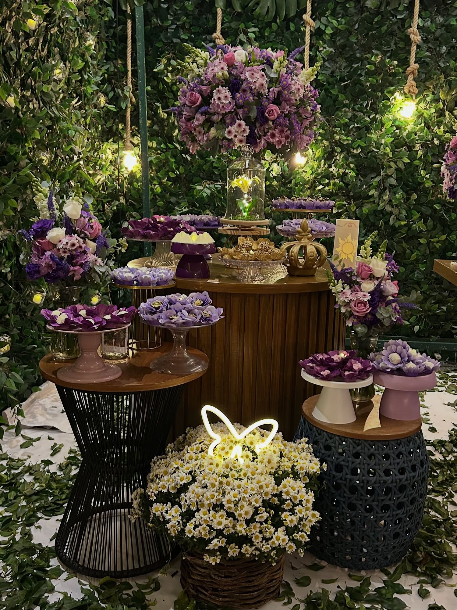 Mesa de doces com arranjos de flores lilás diante de uma parede verde com luzinhas
